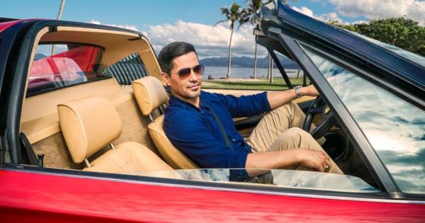 Magnum P.I.sitting back in a co<em></em>nvertible with sunglasses, smiling with his hand on the wheel.