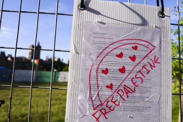 The words Free Palestine and a drawing of a watermelon on a notice of trespass posted on a fence in front of a lawn with tents on it.