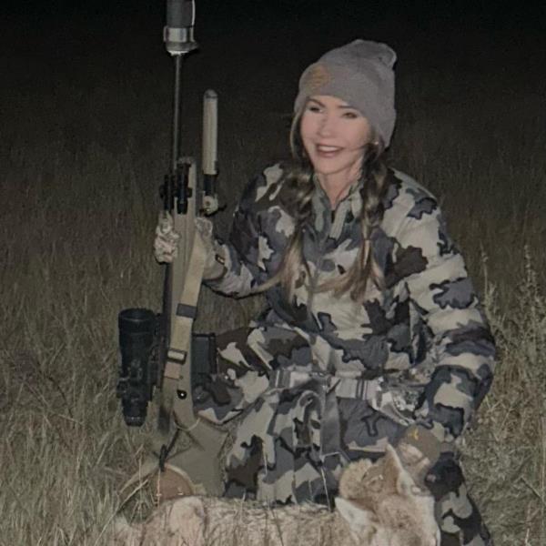 Kristgi Noem wears Army fatigues and bends down over a dead animal while holding a rifle and smiling