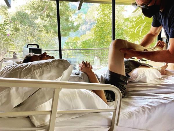 Jeremy Renner lying in bed with a man bending his leg. 