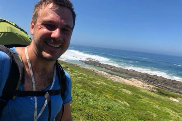 Warwick Tollemache smiles in a selfie on a grassy hill near a beach.