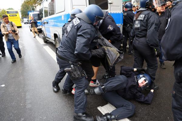 A group of police officers wearing riot gear surround another officer who has fallen down on the street behind a blue and white police van.
