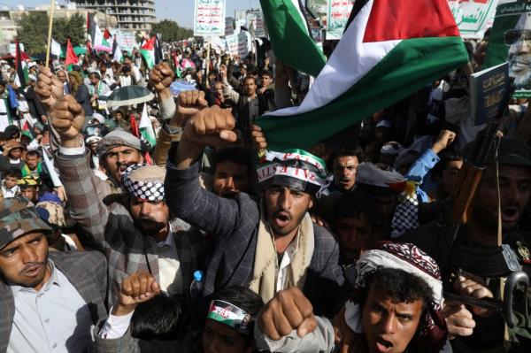 A large group of male demo<em></em>nstrators wearing hats waving their fists at the camera as a large Palestinian flag waves behind them.